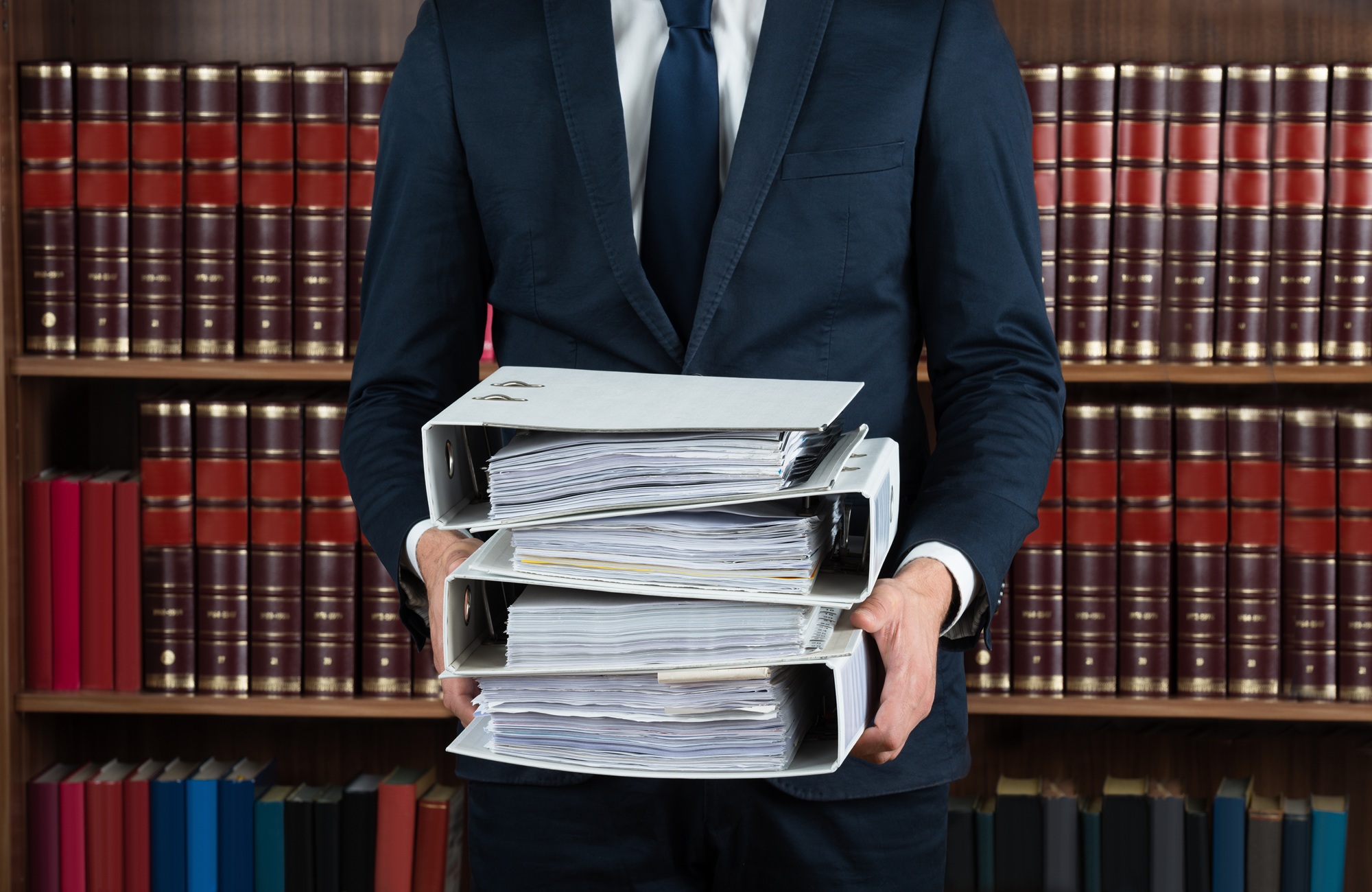 Un avvocato in abito formale tiene in mano diversi raccoglitori pieni di documenti legali davanti a una biblioteca giuridica.